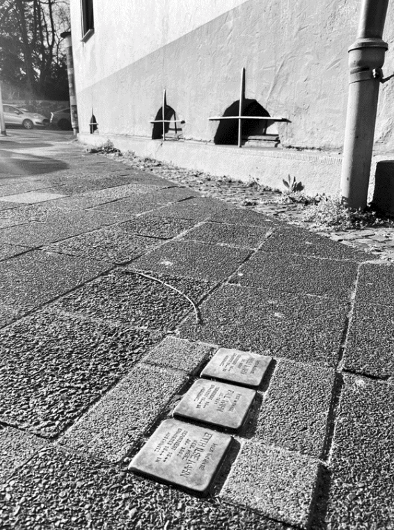Schwarzweißfoto: drei Stolpersteine im Gehweg vor einer Hauswand, Pflaster und Regenrohr im Bild.